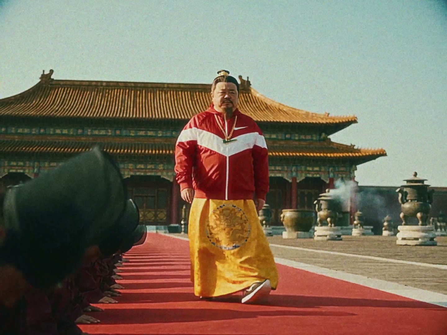 A Chinese emperor in a track jacket walks a red carpet by the palace