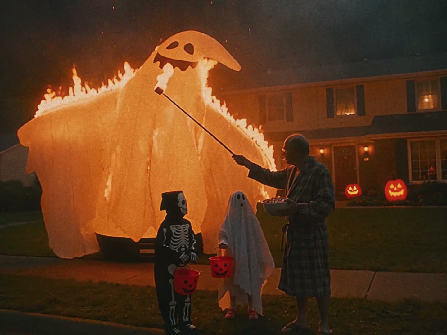A man in a bathrobe toasts a marshmallow over a burning inflatable ghost while kids in costumes wait